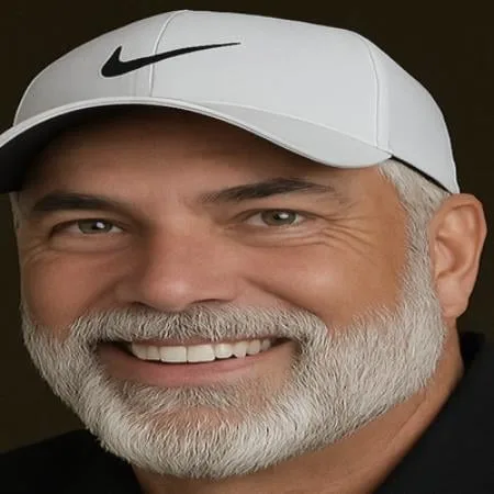 Middle-aged man with gray beard wearing white Nike cap, smiling at camera against dark background