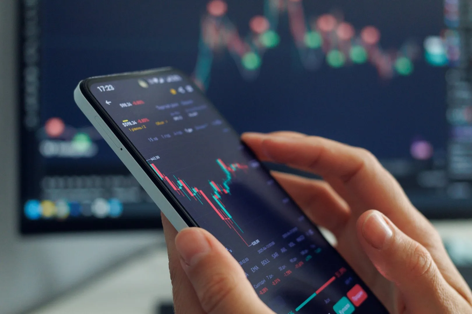 A person's hands hold a smartphone displaying a stock market chart with red and green candles, indicating price fluctuations, against a blurred background showing more financial data on a larger screen, suggesting mobile trading or investment analysis.